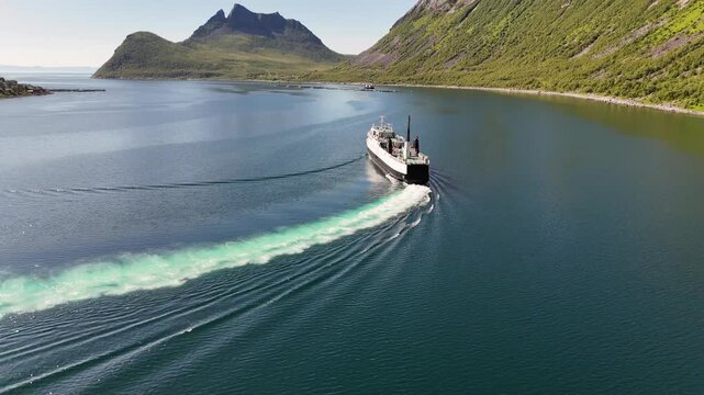 14 july 2025, Gryllefjord, Norway. Aerial, forward drone flight above the water of a fjord, Ferry from Gryllefjord toward Andenes on Lofoten left the harbor, turning in fjord. Village visible, mountai