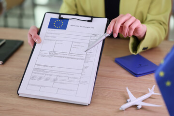 A person reviews a Schengen visa application form while pointing at it with a pen. Clear desk items hint at travel planning, including a small airplane model and a passport.