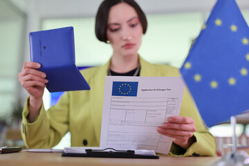 Focused on her work, the woman in a bright outfit carefully examines a Schengen visa application, with European flags in the background, capturing a moment of diligence and purpose.