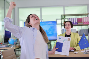 A woman joyfully raises her arm in victory while holding documents in a lively office. A smiling colleague watches, creating a positive atmosphere of achievement and teamwork.