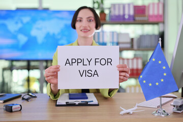 In a vibrant office, a confident woman smiles while holding a sign that says Apply for Visa. Behind her is a modern workspace filled with colorful folders and a digital map.