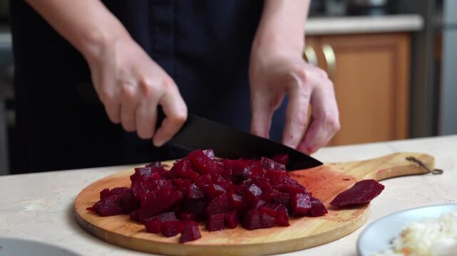 Professional cook carefully cutting boiled beetroot into small cubes with a sharp kitchen knife on a wooden board, preparing ingredients for a healthy salad or traditional dish