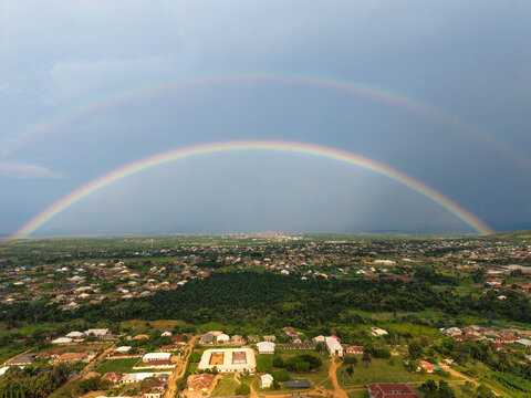 Aerial view of a vibrant double rainbow arcing across the sky over a dense landscape of buildings and lush greenery, Ilesa, Osun, Nigeria.