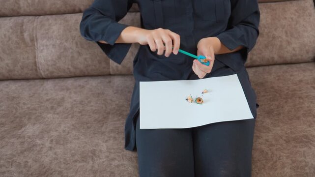 Close-up of female hands sharpening a graphite pencil with a blue sharpener, with shavings and broken tips falling onto a white paper sheet, representing preparation for drawing or studying