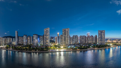 Fototapeta premium Modern residential skyscrapers illuminated at night along a riverbank under a deep blue sky.