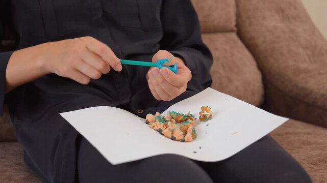 Close up view of a person's hands holding a pencil sharpener and carefully sharpening a turquoise colored pencil, with the wood and graphite shavings collecting on a piece of white paper