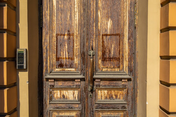 Pair of aged wooden doors with rich, textured grain and warm brown hues stands beside a beige wall. The rustic charm of the weathered wood contrasts with the sleek, metallic intercom panel mounted to 