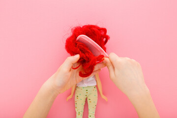 Little child girl hands holding plastic comb and combing doll red messy hair on light pink table background. Pastel color. Closeup. Point of view shot. Top down view.