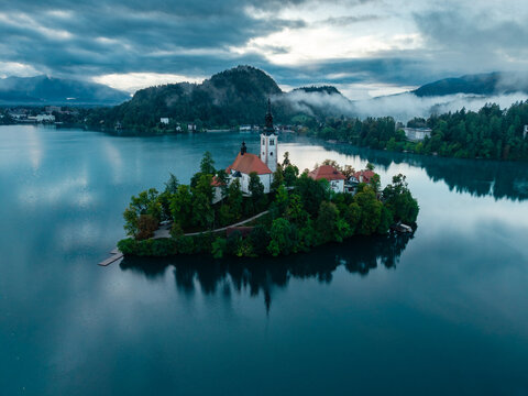 Aerial view of the iconic church on Bled Island amidst the tranquil lake waters reflecting the moody sky, framed by misty mountains, Bled, Radovljica, Slovenia.