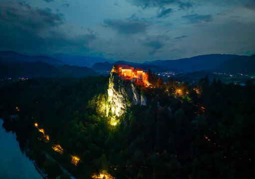 Aerial view of Bled Castle perched atop a steep rock, illuminated against the dark, forested landscape, a beacon of light under the twilight sky, Bled, Radovljica, Slovenia.