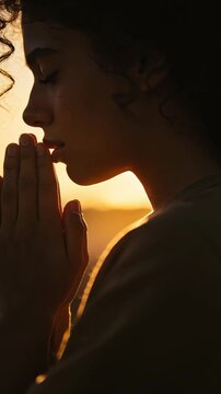 Young woman praying at sunset in side profile with hands clasped, spiritual faith and meditation with warm golden light.