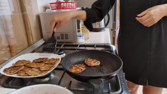 Close-up view of a woman's hands using a spatula to fry traditional golden potato pancakes, or draniki, in hot oil on a gas stove, preparing a delicious homemade meal in the kitchen