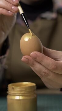 Female hands coloring eggs with golden paint on blurred garland lights on background. Easter, tradition, holidays and people