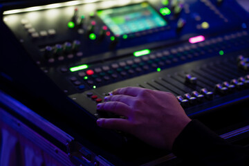 Fototapeta premium Hand on digital audio mixing console in road case, adjusting faders and backlit buttons for live event sound control under blue stage lighting