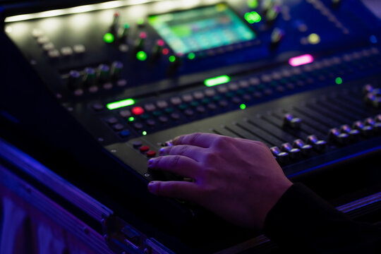 Hand on digital audio mixing console in road case, adjusting faders and backlit buttons for live event sound control under blue stage lighting