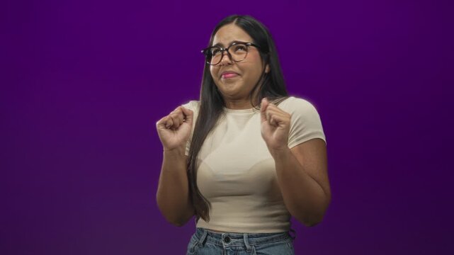 Young woman with glasses and hands raised, recoiling with a disgusted facial grimace, wearing beige tee and jeans in a purple studio; disgust avoidance.