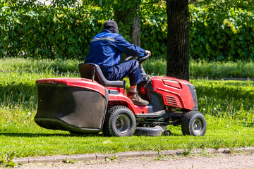Person in blue work uniform meticulously mows lush green grass using a red riding lawn mower. The scene captures everyday garden maintenance amidst a serene, tree-lined area, blending labour with the 