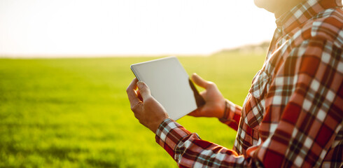 A man stands in a green field at sunset, holding a tablet. The sunlight creates a warm glow. The man is wearing a plaid shirt and appears to be staring intently at the device. © maxbelchenko