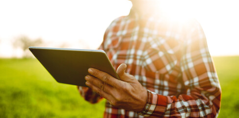 A man stands in a green field at sunset, holding a tablet. The sunlight creates a warm glow. The...