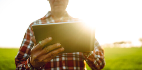 A man stands in a green field at sunset, holding a tablet. The sunlight creates a warm glow. The man is wearing a plaid shirt and appears to be staring intently at the device. © maxbelchenko