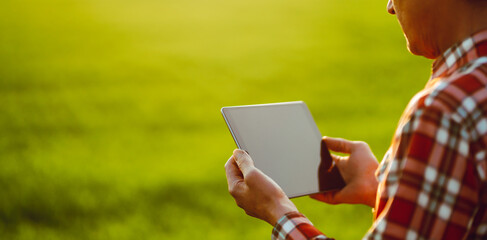 A man stands in a green field at sunset, holding a tablet. The sunlight creates a warm glow. The man is wearing a plaid shirt and appears to be staring intently at the device. © maxbelchenko