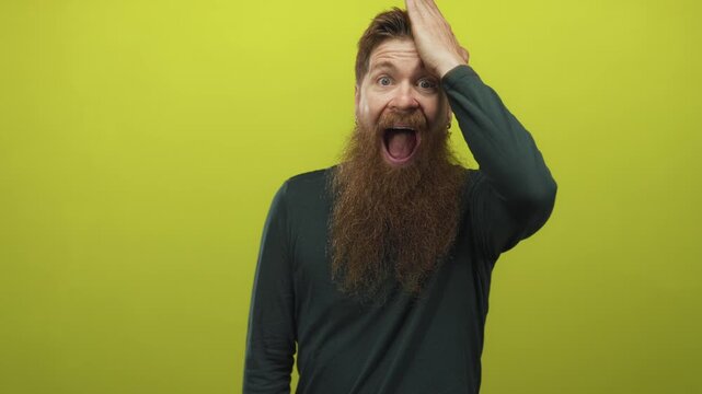 Young caucasian man with long beard touching forehead against bright green studio backdrop; surprise.
