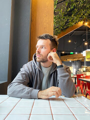 Young Caucasian man with short hair wearing a gray hoodie rests his chin on his hand while sitting at a table in a modern cafe with indoor plants