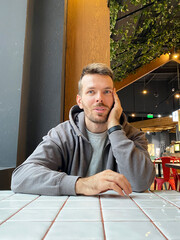 Young Caucasian man with short hair wearing a gray hoodie rests his chin on his hand while sitting at a table in a modern cafe with indoor plants