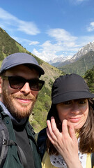Couple wearing sunglasses and caps pose for a selfie in a mountainous landscape with green hills and snow-capped peaks under a blue sky