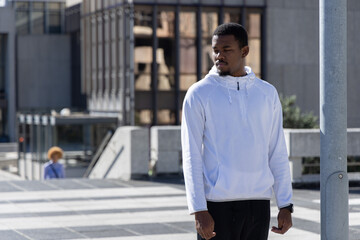 Fototapeta premium African American man standing near metal light pole on tiled plaza wearing white hoodie, copy space