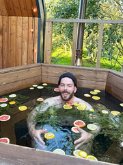 A man in a wooden tub filled with citrus fruits and fir branches enjoys a relaxing moment surrounded by greenery on the veranda of a country house in summer. Outdoor bathroom