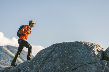 African American man hiking up a mountain with a backpack in an orange coat with ski area trails off in the distance in Vermont, New England, USA.