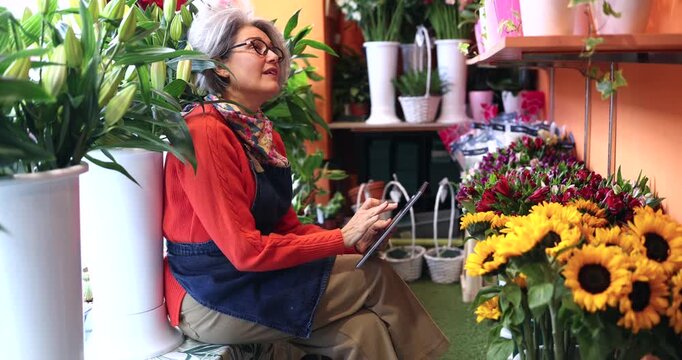 Experienced senior female florist sitting among flowers in her shop, using a digital tablet to manage orders and inventory for her small business, smiling with satisfaction