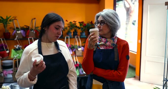 Cheerful female florists of different ages wearing aprons are enjoying a relaxing coffee break together and chatting inside a flower shop. Colleagues sharing a friendly moment during work hours