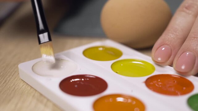 Close-up of woman hands takes white paint for coloring easter eggs with painting brush. Holidays, tradition and people