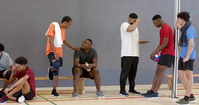 Diverse male volleyball team resting in gym, coach scrolling tablet then guiding return to play