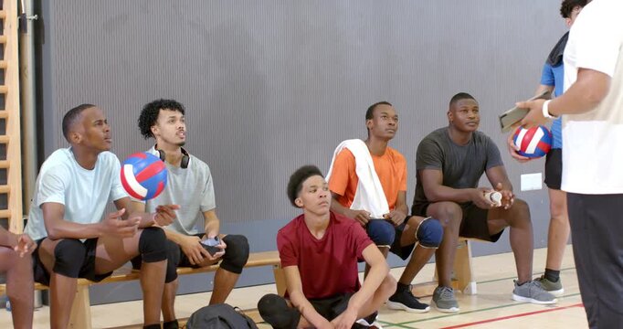 Coach tapping clipboard and pointing, guiding male teammates with volleyballs leaning on sideline