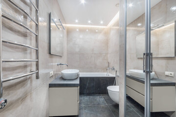 A bathroom with a bathtub, washbasin, and toilet. The walls are soft and light colored. The modern design includes a towel rack and mirrors reflecting the space