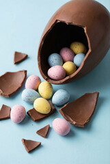 Broken chocolate easter egg spilling out colorful speckled candy eggs on a light blue background.