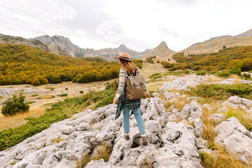 Fototapeta premium View from behind. A hiker with a backpack and hat walks along a rocky mountain trail. The young woman enjoys the mountain scenery and feels a sense of freedom. Concept of hiking, nature, and travel.