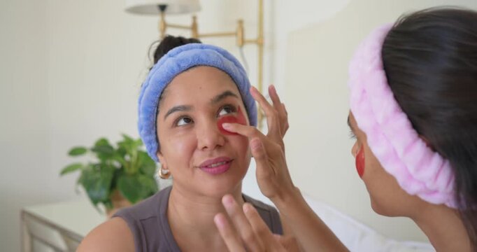 Female friends sitting on bed, right friend mirroring own patch, placing red patch under left eye