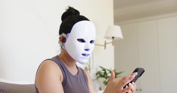 Asian woman is lifting phone, tapping screen, and adjusting LED mask on couch during home skincare