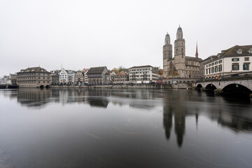 Obraz premium Long-exposure view of Zurich’s old town on a cold, foggy winter day, muted tones and a gloomy mood, with the smooth Limmat River in the foreground near the cathedral.