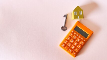 An orange calculator, a small yellow wooden house, and a vintage metal key are arranged on a pale pink surface, representing the financial calculations involved in home ownership and real estate.