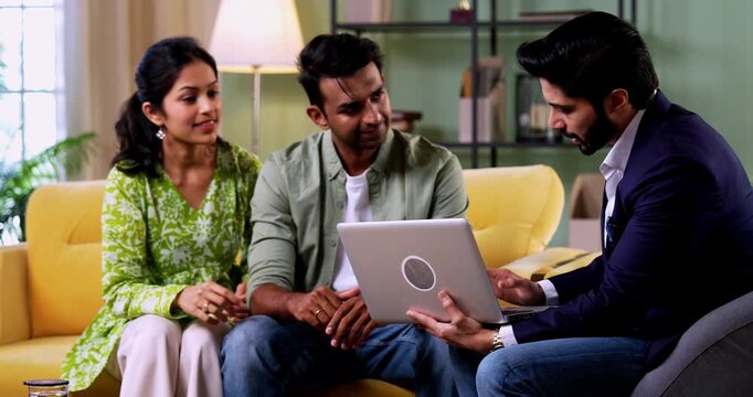 Indian couple meeting agent at home with laptop during investment discussion with bank representative reviewing financial plans while sitting on sofa in cozy modern living room, home consultation