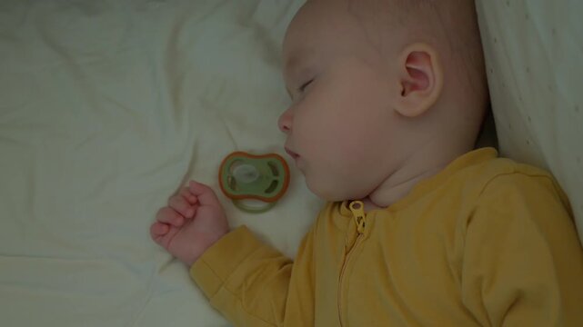 caucasian baby sleeping beside pacifier, dreaming sleeper in yellow onesie on soft pillow with gentle nightlight glow, serene nap routine, tranquil nursery ambiance