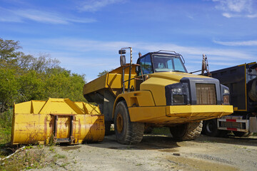 Obraz premium Dump truck with attachment. Yellow articulated dump truck and yellow attachment container parked on gravel near trees and other trucks.
