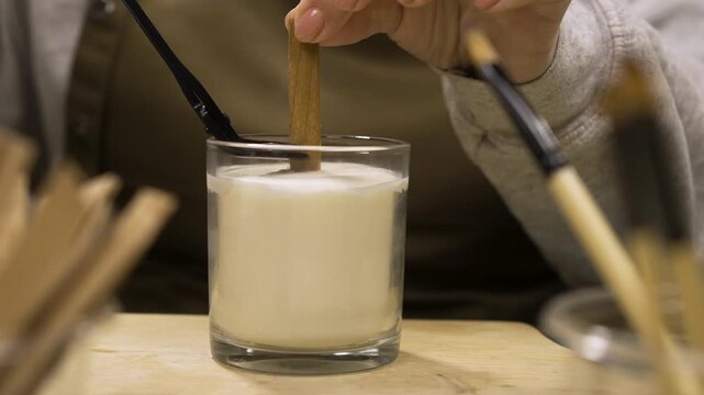 Close-up of female hands cutting wooden wick of homemade candle with professional scissors at table indoors