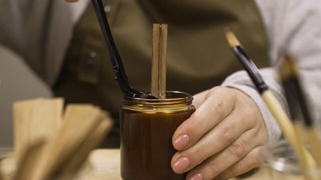 Close-up of female hands cutting wooden wick of homemade candle with professional scissors at table indoors
