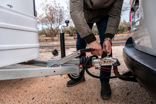 Man attaching a small white trailer to a vehicle, preparing for travel. Connection involves hands operating the hitch mechanism for secure towing